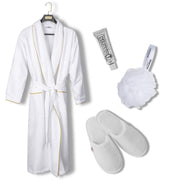 A white bathrobe with gold trim hanging on a black hanger, accompanied by a silver tube of body wash, a white loofah bath sponge, and a pair of white slip-on slippers on a plain white background. Shop Meriton Suites.