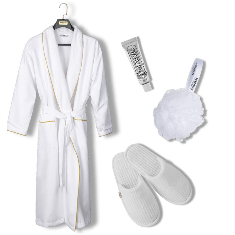 A white bathrobe with gold trim hanging on a black hanger, accompanied by a silver tube of body wash, a white loofah bath sponge, and a pair of white slip-on slippers on a plain white background. Shop Meriton Suites.