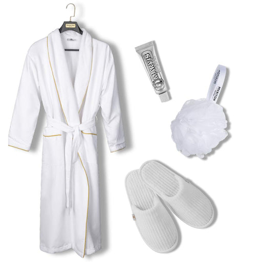 A white bathrobe with gold trim hanging on a black hanger, accompanied by a silver tube of body wash, a white loofah bath sponge, and a pair of white slip-on slippers on a plain white background. Shop Meriton Suites.