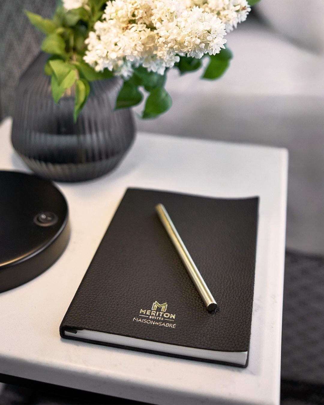 Black leather notebook with gold pen and Meriton Suites logo on a desk near a lamp and plant.