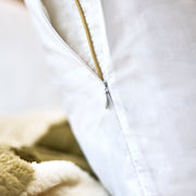 A close-up of a white pillowcase with a gold zipper, resting on a soft beige and olive green sherpa blanket.