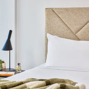 A modern bedroom scene featuring a bed with white linens, a textured beige geometric headboard, a black conical bedside lamp, and a window overlooking a waterfront landscape.