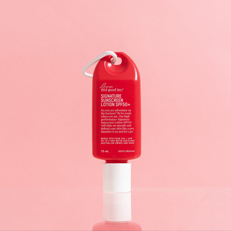 A red and white bottle of WAFGI Signature Sunscreen Lotion SPF 50+ with a white carabiner clip, centered against a solid pink background.