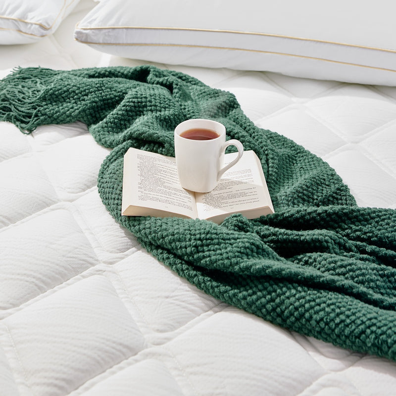 Green knitted blanket draped over a bed with an open book and a mug of tea.