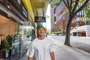 Man wearing a green helmet standing on a city street with a building and tree in the background. Shop Meriton Suites.