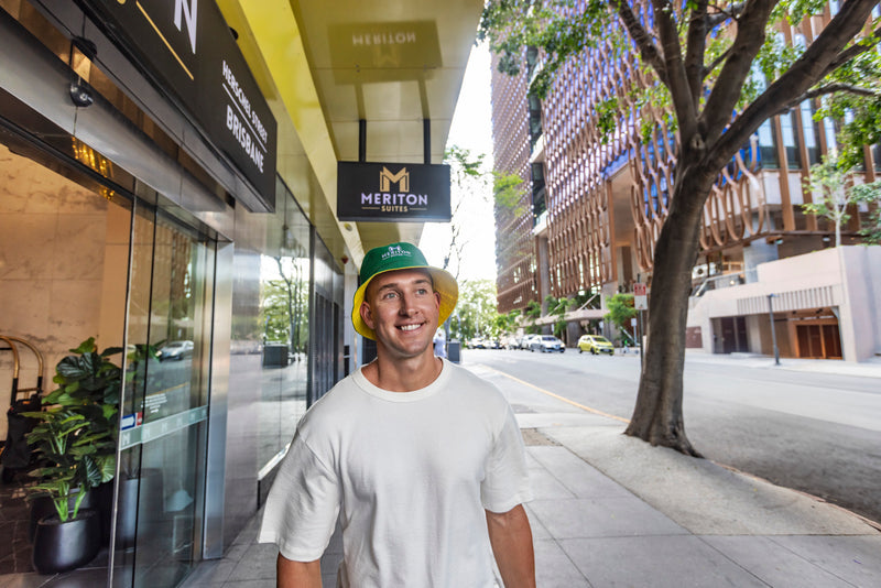 Man wearing a green helmet standing on a city street with a building and tree in the background. Shop Meriton Suites.