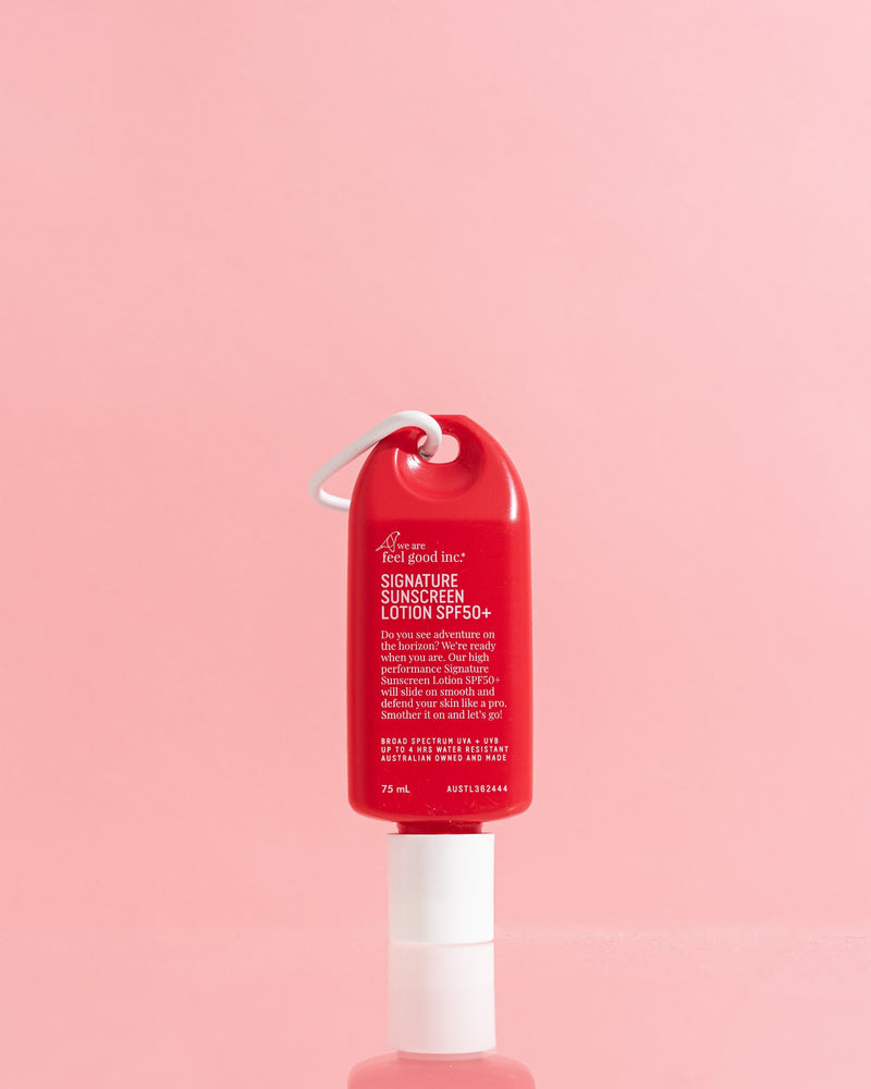A red bottle of Sunscreen Lotion SPF 30 with a white pump dispenser and a white plastic carabiner loop, set against a solid pink background. Shop Meriton Suites. We are feel good inc. spf sunscreen.