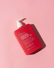 A red rectangular pump bottle of Signature Sunscreen Lotion SPF 50+ with a white pump, photographed on a solid pink background with a sharp shadow. Shop Meriton Suites. We are feel good inc. spf sunscreen.