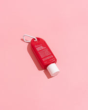 A small red bottle with a white cap and white loop, photographed diagonally on a solid pink background.  Shop Meriton Suites. We are feel good inc. spf sunscreen.