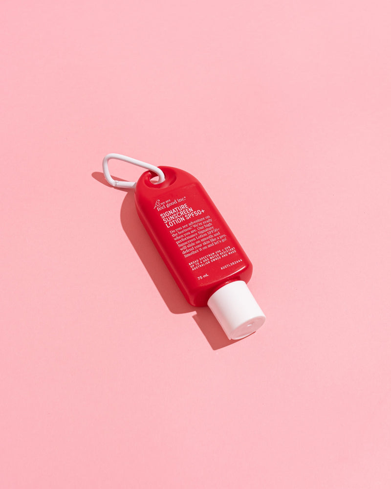 A small red bottle with a white cap and white loop, photographed diagonally on a solid pink background.  Shop Meriton Suites. We are feel good inc. spf sunscreen.