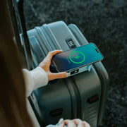 A close-up lifestyle shot of a person's hand holding a smartphone over a gray hard-shell suitcase, demonstrating wireless charging. The phone screen displays a green charging icon. Shop Meriton Suites SnapWirless